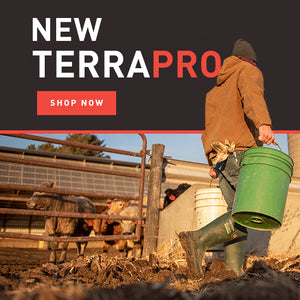 Man holding a green bucket whilst walking and wearing cold weather clothing and a pair of green Dunlop TerraPro wellingtons on a farm with buildings and livestock in the background. Text reads 'NEW TERRAPRO. SHOP NOW'
